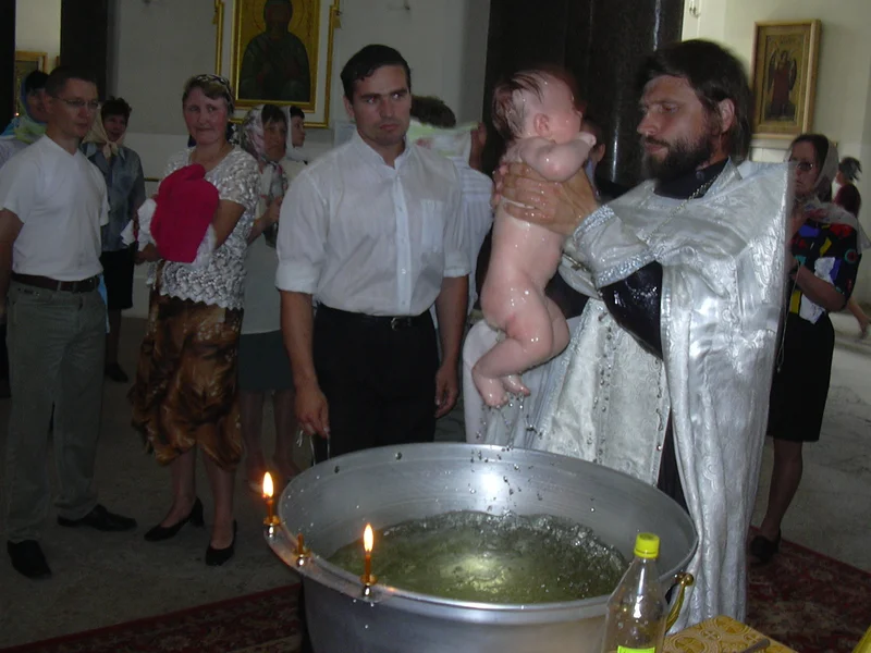 Священник держит младенца над купелью; рядом стоят родные и восприемники