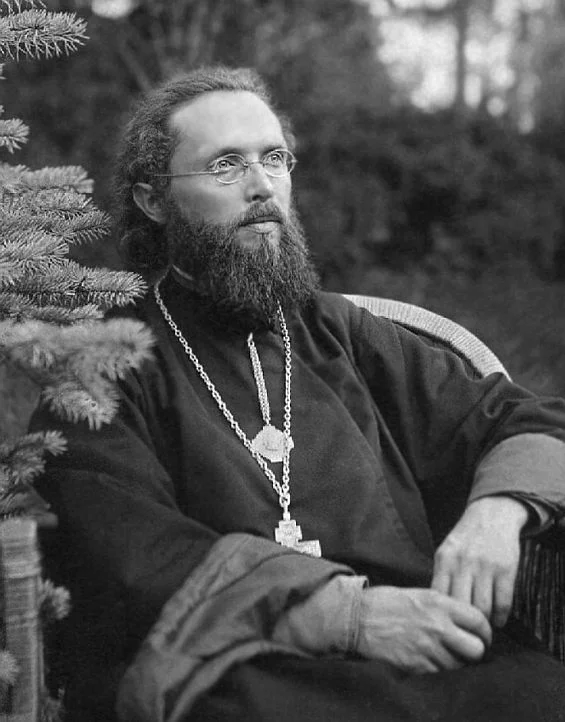 Чёрно-белая фотография молодого священника в подряснике с наперсным крестом, на фоне ели